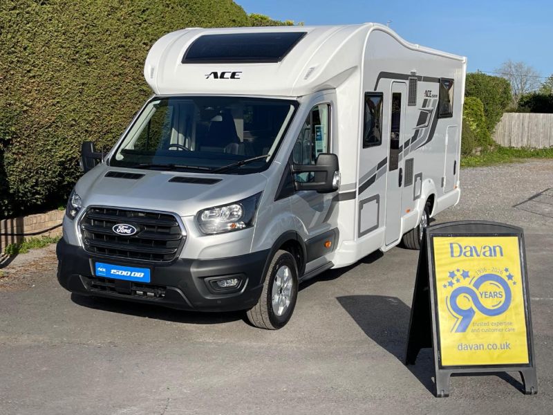 White ACE motorhome (RV) parked on a paved lot beside a yellow Davan signboard showing 90 years preserved celebration.
